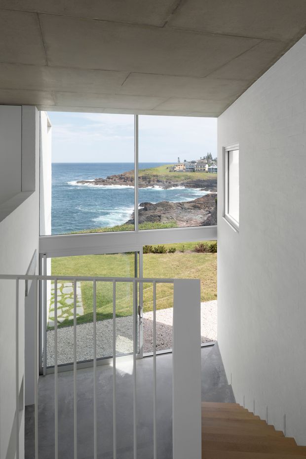 A minimalist interior stairway descending beside large floor-to-ceiling glazing, with a white painted wall, exposed concrete ceiling and grey polished concrete landing. White vertical balustrades frame expansive views of a rugged ocean coastline, lawn and nearby coastal buildings. Timber stair treads add warmth to the otherwise restrained monochrome interior.