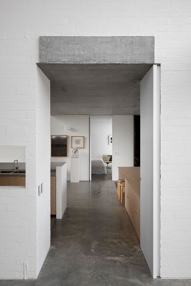 A minimalist interior view through a wide doorway, showing a polished concrete hallway beneath a substantial exposed concrete lintel or soffit. White-painted brick walls frame the opening, with a glimpse of a kitchen and sink through a wall opening on the left, fixed light-wood joinery and stools on the right, and a bedroom visible at the rear. The composition emphasizes strong geometric lines, restrained materials, and neutral tones.
