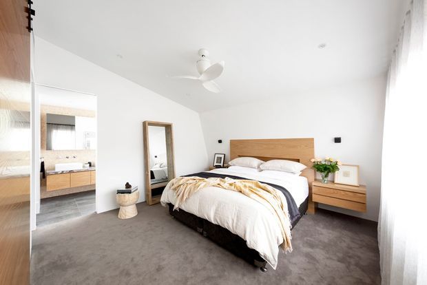 Bright contemporary bedroom with a large upholstered bed, timber headboard and matching floating bedside cabinetry. The room has white walls and ceiling, grey carpet, sheer full-height curtains, a ceiling fan, and a full-length mirror. An ensuite bathroom is visible through the open doorway at left.
