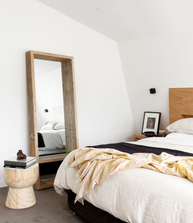 Bright, minimalist bedroom with white walls and ceiling, a low bed dressed in white linen with a dark throw and cream blanket, and a simple timber headboard. A large rustic timber-framed floor mirror leans against the wall, reflecting the bed. A small rounded timber side table holds books and a decorative object, while a bedside table displays framed artwork and a bowl. The room has a calm, neutral palette with natural wood accents and soft daylight.