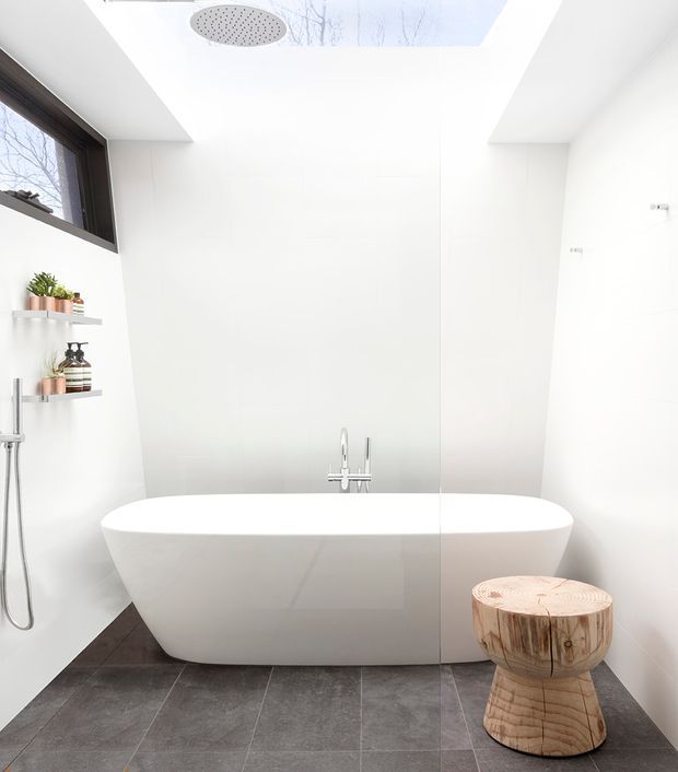 Bright, minimalist bathroom with a freestanding white bathtub positioned beneath a large skylight. The room has white wall surfaces, dark grey floor tiles, a wall-mounted shower with recessed shelving and small plants, a dark-framed window, and a sculptural natural timber stool beside the bath.