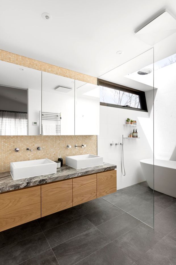 Bright contemporary bathroom featuring a floating timber vanity with two white vessel basins, a dark veined stone benchtop, mirrored wall cabinetry, and a warm hexagonal mosaic tile splashback. A frameless glass shower with recessed shelving and a freestanding white bathtub occupy the right side. Large grey floor tiles, white walls, skylights, and a high horizontal window create an airy, refined space.