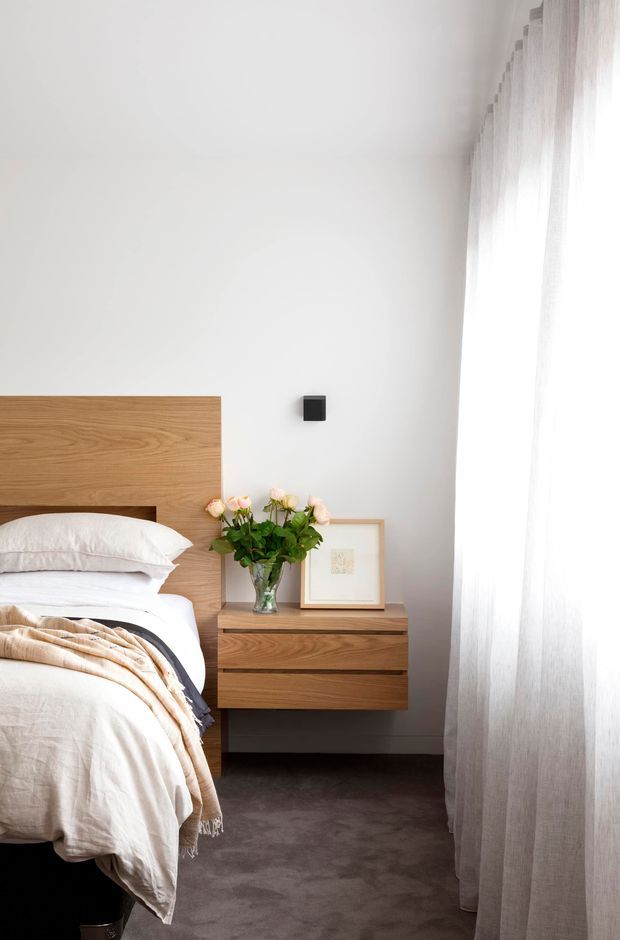 A bright, minimalist bedroom vignette with a partially visible bed, a simple timber headboard, and a floating timber bedside cabinet. White bedding, sheer full-height curtains, a vase of flowers, and framed artwork create a soft, understated atmosphere. The room has a neutral palette and abundant natural light.