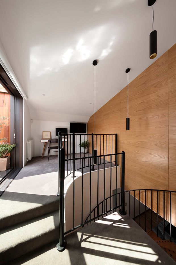 A bright modern stairway and landing with a curved stair descending beside a tall timber-panelled wall. Black metal balustrades and slim black pendant lights contrast with the warm wood, while a large glazed opening brings in strong sunlight. A small desk and television are visible on the upper landing.