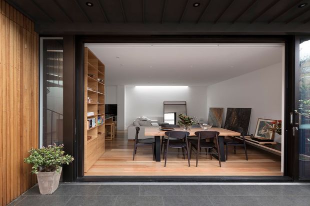 View from an outdoor terrace through wide black-framed sliding glass doors into an open-plan dining and living area. The interior features a timber dining table with dark chairs, pale timber flooring, built-in timber shelving, a light sofa, and artwork. The exterior threshold has dark grey tiled paving, while the surrounding facade and soffit use warm vertical timber cladding and timber boards. The space has a refined contemporary character with strong indoor-outdoor connection.