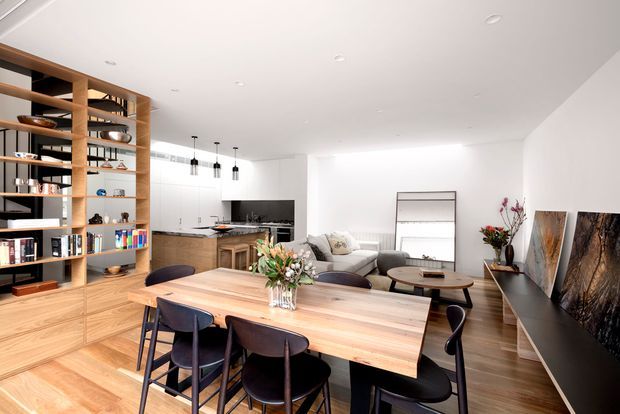 Bright open-plan contemporary interior combining a dining area, kitchen and living room. A large light timber dining table with dark upholstered chairs occupies the foreground, while a timber built-in shelving and cabinetry wall lines the left side. The kitchen has a dark benchtop and timber joinery, and the rear seating area includes a light sofa and round coffee table. White walls and ceiling, recessed lighting, pendant lights, timber flooring, and restrained decorative styling create a clean, warm, refined atmosphere.