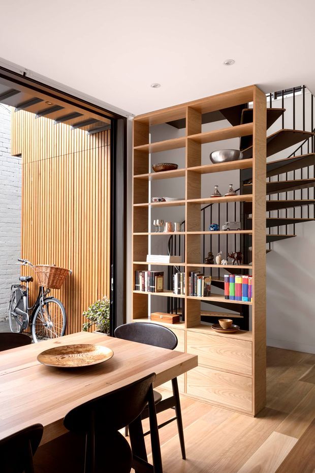 Bright contemporary dining area with a substantial built-in timber shelving and storage wall, displaying books and decorative objects. A timber dining table with dark chairs occupies the foreground. To the left, a large glazed opening connects to an enclosed courtyard or lightwell with vertical timber screening, white brickwork, and a bicycle. A slim open-riser staircase with dark metal framing is visible behind the shelving.