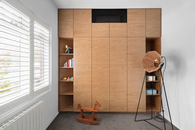 A bright, compact interior workspace or study with a full-height built-in timber storage and cabinetry wall. The central doors conceal storage, while open shelving on both sides displays books and small objects. A sculptural floor lamp and wooden rocking horse sit on a dark polished concrete or dark grey floor. White plantation shutters and a radiator line the large window on the left. The design is clean and contemporary, with warm timber joinery contrasting against white walls and a dark floor.