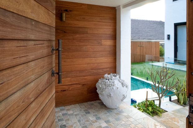 A timber-clad entryway opens toward a courtyard pool. The space features a large horizontal-plank timber door and wall, dark metal door hardware, a mosaic-tiled floor, white rendered columns and walls, and a sculptural white planter. Through the opening is a turquoise swimming pool, landscaped courtyard, and neighbouring timber fencing.