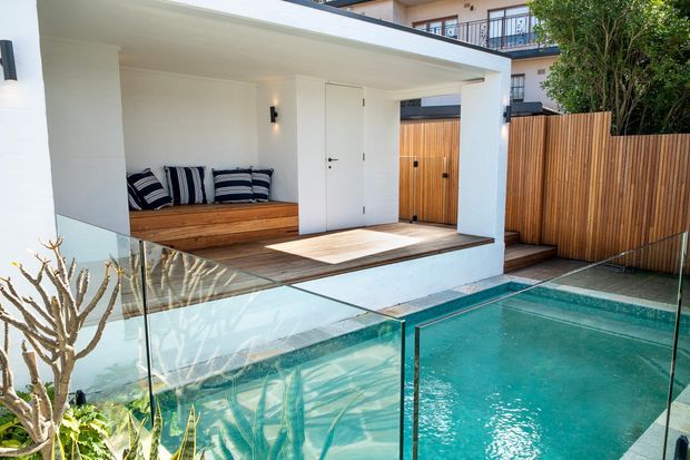 A contemporary pool courtyard with a covered outdoor lounge or entry area. The space features a turquoise swimming pool enclosed by frameless glass balustrades, pale tiled paving, a timber built-in bench with striped cushions, white rendered walls, a timber privacy fence, and a timber-clad gate.