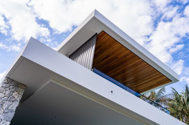 Low-angle exterior view of a contemporary multi-level residence with crisp white geometric volumes, a large projecting roof or balcony canopy, warm timber soffit lining, blue-tinted glass balustrading, vertical metal screening, and a natural stone wall section. Palm vegetation and a partly cloudy sky surround the building.