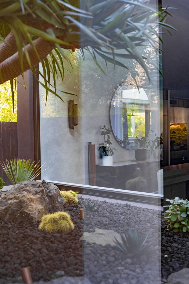 A view through floor-to-ceiling glazing into a refined interior, with strong reflections of tropical foliage over a minimalist courtyard garden. The interior features a pale textured wall, dark timber or charcoal cabinetry, a rounded wall mirror, potted plants, and sculptural decorative elements. Outside, dark gravel, rocks, grasses, and small rounded cacti create a Japanese-inspired xeriscaped garden. The image is calm and layered, combining indoor and outdoor views through reflective glass.