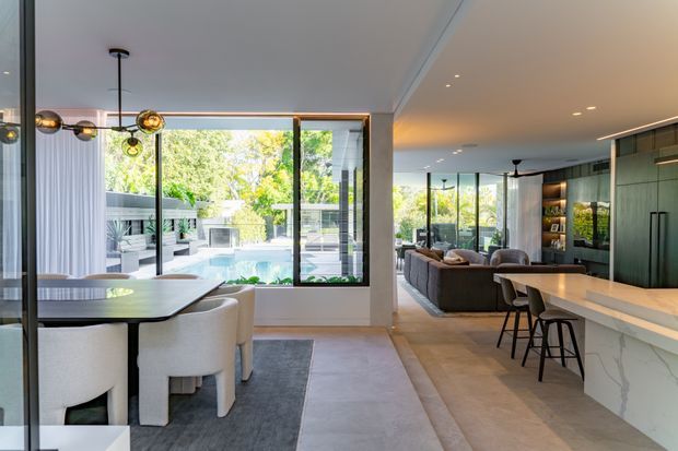 Open-plan contemporary residential interior combining a dining area, kitchen and living room. The space features expansive black-framed glazing overlooking a landscaped pool courtyard, a long stone kitchen island with bar seating, dark green built-in cabinetry, pale stone-look flooring, and a sculptural cluster pendant above the dining table. The interior is bright, refined and connected to the outdoors.