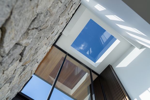 Upward-looking architectural interior view showing a double-height white ceiling with a large blue abstract artwork or skylight-like feature, strong daylight, exposed natural stone walling, timber surfaces, black-framed glazing, and dark vertical timber slats. The composition emphasizes the relationship between the stone wall, overhead void, and modern structural elements.