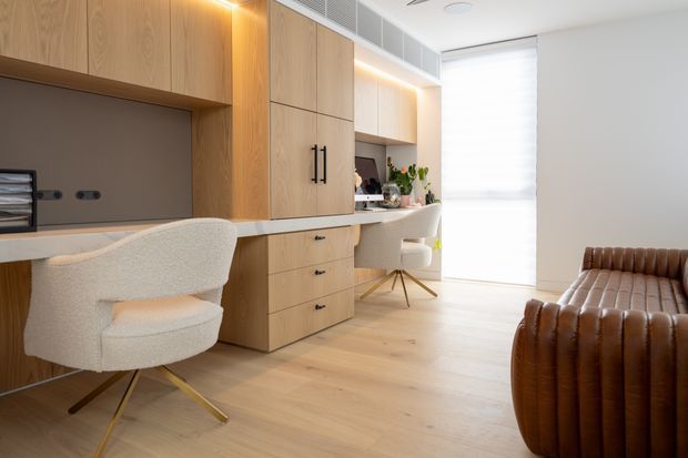 Bright, refined home office with custom light-wood cabinetry and integrated desks along one wall. Two upholstered work chairs sit at the desks, with a small vanity or secondary workspace near the window. The room has pale timber flooring, warm concealed joinery lighting, white walls, a large diffused window, and a brown ribbed leather sofa or lounge chair in the foreground.