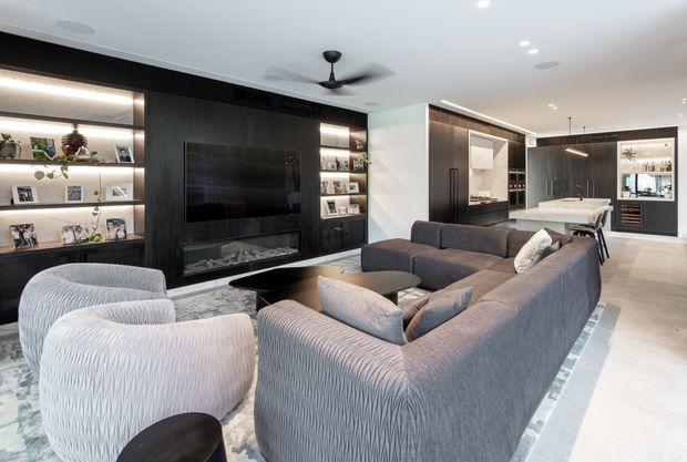 A spacious open-plan contemporary living room with a large grey sectional sofa, two textured lounge chairs, a dark oval coffee table, and a patterned area rug. A full-height dark timber media wall incorporates a television, linear fireplace, open display shelving, and decorative lighting. The room opens toward a dining area and a dark-toned kitchen with an island in the background. Recessed ceiling lights, a ceiling fan, pale flooring, and clean-lined built-in joinery create a refined, modern atmosphere.