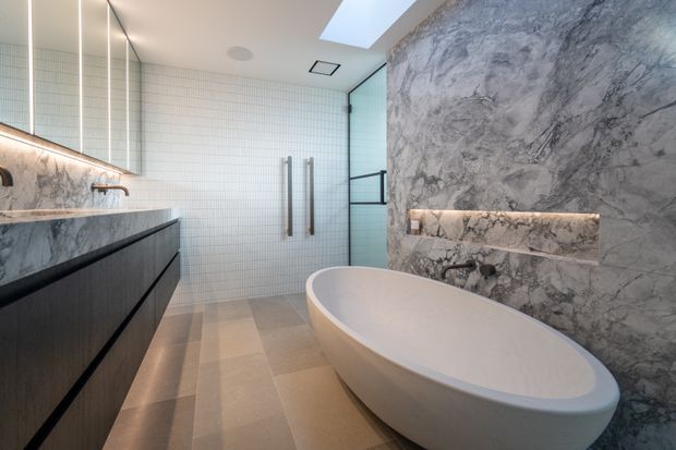 Contemporary bathroom featuring a freestanding oval bathtub, double vanity with dark timber-look cabinetry, marble-look wall surfaces, a small white grid-tile wall, frameless glass shower enclosure, recessed wall niche with integrated lighting, skylight, and linear illuminated mirror lighting. The palette is predominantly warm beige, white, grey, and dark wood, creating a refined spa-like atmosphere.