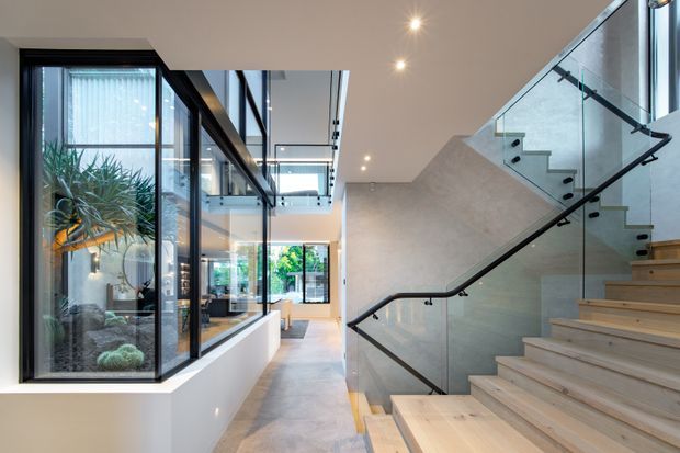 A bright contemporary interior hallway and stairway with a dramatic glazed internal courtyard or atrium on the left. Pale timber stairs rise beside a grey textured wall, with clear glass balustrades and black metal handrails. Full-height black-framed glazing, polished light flooring, recessed downlights, and indoor planting create an airy, refined circulation space.