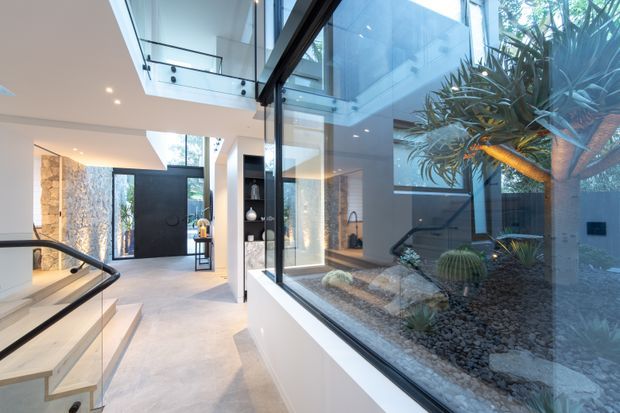 A bright contemporary interior entry hall with a sculptural stair and glass balustrade on the left, a large black pivot-style entry door at the end, and a full-height glazed internal courtyard containing rocks, cacti, and a mature architectural plant. White walls, pale flooring, black-framed glazing, recessed lighting, and a visible upper-level void create an airy, gallery-like space.