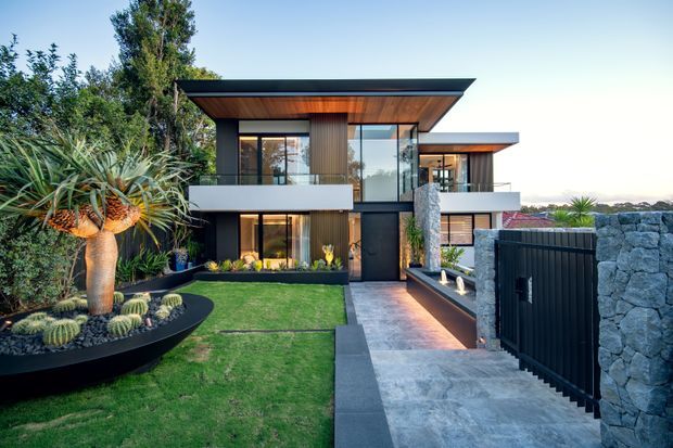Contemporary multi-level residence with bold projecting flat rooflines, extensive floor-to-ceiling glazing, dark metal framing, warm vertical timber cladding, and pale balcony bands. The landscaped front garden includes sculptural planting and cacti, while a paved entry path leads past a dark slatted gate and rough grey stone boundary wall.