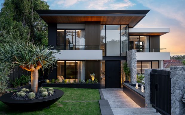 Contemporary two-storey residence viewed from the landscaped front garden, featuring strong horizontal flat roof planes, dark cladding and window frames, extensive floor-to-ceiling glazing, warm timber soffits, white rendered volumes, and a prominent textured stone entry wall. The approach includes sculptural desert planting, lawn, paved paths, and a dark metal gate and fence.