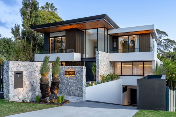 Contemporary multi-level residence viewed from the street, with a strongly horizontal flat-roofed composition, extensive glazing, dark vertical cladding, white rendered walls, and rough natural-stone retaining and boundary walls. A recessed garage is accessed by a sloping driveway, while landscaped planting softens the facade.