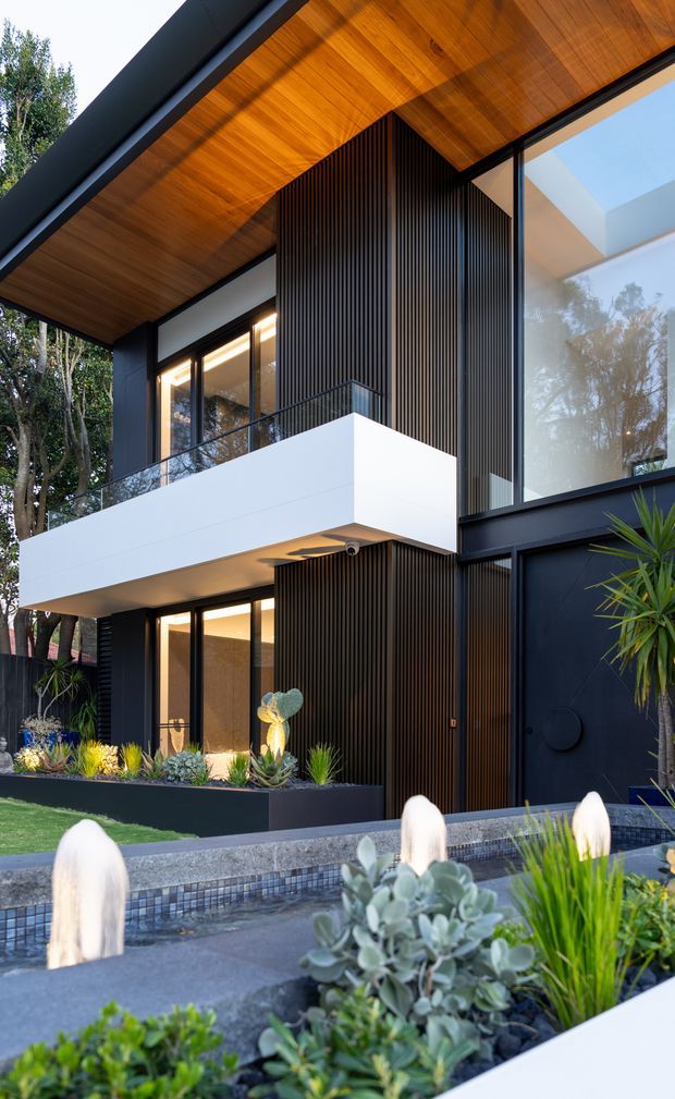 A contemporary multi-storey house facade viewed from the landscaped front garden. The building features dark cladding, extensive black-framed glazing, a projecting white balcony, vertical dark timber-look battens, and a warm timber-lined pitched roof soffit. Layered planting, raised planters, and a narrow water feature occupy the foreground.