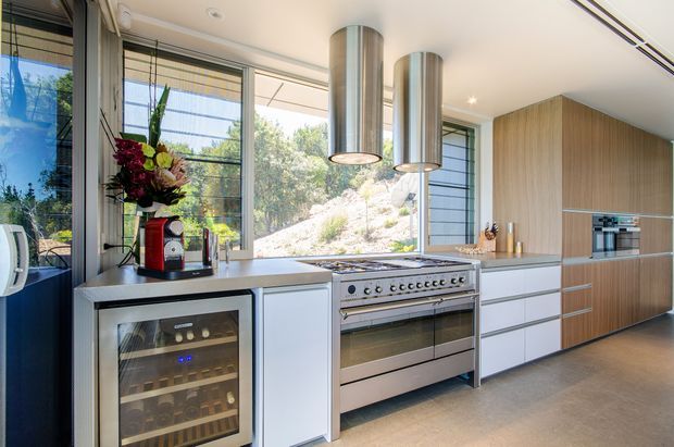 Bright contemporary kitchen with extensive floor-to-ceiling glazing overlooking a rocky landscaped setting. The room features pale flat-panel cabinetry, a long dark benchtop, a large stainless-steel range cooker, integrated appliances, cylindrical stainless-steel pendant rangehoods, and a built-in wine refrigerator.