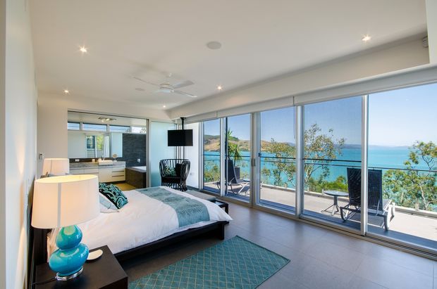 Bright contemporary bedroom with a large bed, bedside lamps, a ceiling fan, and full-height sliding glass doors opening to a balcony or terrace. The glazing provides expansive views over vivid blue water and a hilly coastal landscape. The room uses pale walls, dark timber-toned flooring, and restrained modern furnishings.