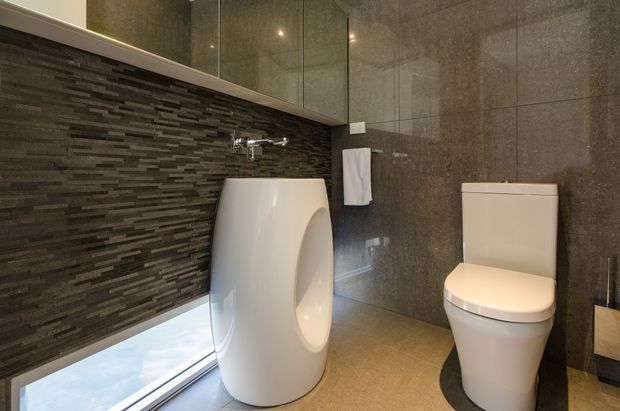 Compact contemporary bathroom featuring a sculptural freestanding white basin, wall-mounted tapware, white toilet, dark textured mosaic-tile feature wall, large grey stone-look wall tiles, mirrored cabinetry, and a narrow low window.