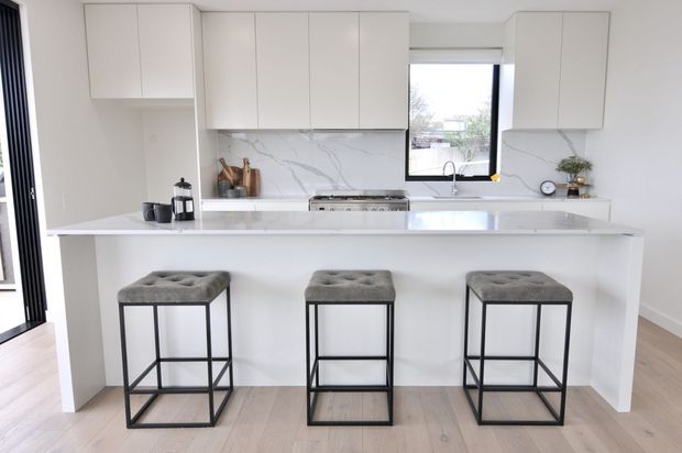 Bright contemporary white kitchen with a large island breakfast bar seating three. The space features handleless white cabinetry, a white marble-look splashback and benchtop, stainless steel cooking appliances, black-framed window and doors, and pale timber flooring. Grey upholstered stools with black metal frames provide contrast.