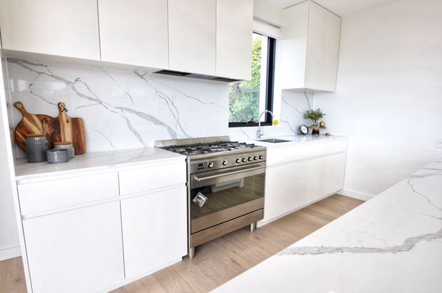 Bright contemporary white kitchen with flat-panel cabinetry, a stainless steel range, white marble-look veined splashback and matching island benchtop, light timber flooring, and a large window above the sink.