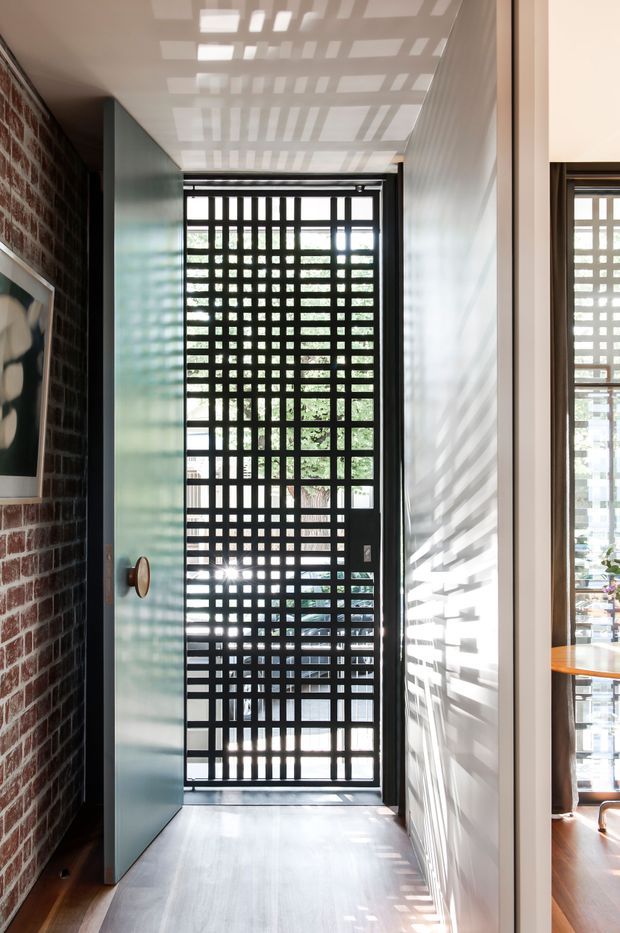 A narrow, light-filled entryway with a black metal grid security door and patterned daylight casting strong geometric shadows across the white ceiling and wall. The left wall is exposed red brick with framed artwork, while a pale green-blue door stands open. A glimpse of a warm timber-floored room is visible through the opening on the right.