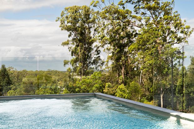An elevated outdoor swimming pool with clear glass balustrades, surrounded by mature trees and dense greenery. The pool edge frames a broad view across a wooded landscape beneath a partly cloudy sky.