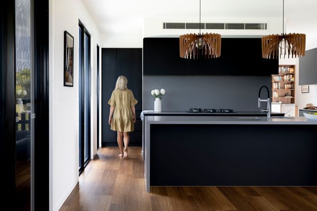 A contemporary open-plan kitchen viewed from the living area, featuring a large black island with a light stone benchtop, matching dark cabinetry, integrated black splashback, timber flooring, and a black-framed glazed opening. Two suspended timber pendant lights hang above the island, while open shelving and a small dining or living area are visible beyond. The space is bright, refined, and strongly contrasted, with a hallway and figure in the background.