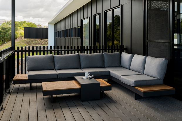 A contemporary outdoor lounge on a raised timber deck, furnished with a large charcoal-grey L-shaped sectional sofa and low timber-and-black tables. The space is enclosed by black vertical balustrading and framed by a dark, vertically panelled building facade with tall glazing. Trees and neighbouring buildings are visible beyond the deck.