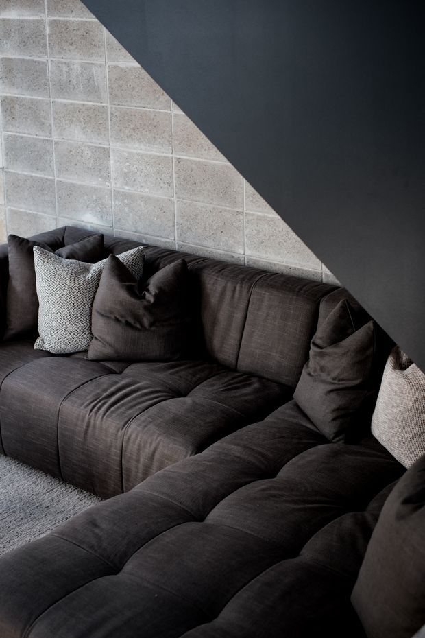 Close-up interior view of a dark charcoal modular sectional sofa with textured cushions, set against a grey tiled feature wall and a dramatic dark angular wall or ceiling plane. The space has a moody, contemporary aesthetic with layered charcoal and grey tones.