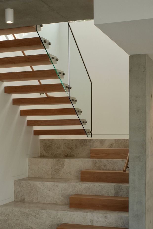 Interior architectural photograph of a contemporary stairway combining floating warm timber treads with pale natural stone steps, a clear glass balustrade, slim dark metal handrail, white walls, and exposed concrete ceiling and structural column. The stair turns at a landing and is illuminated by simple cylindrical ceiling lighting.