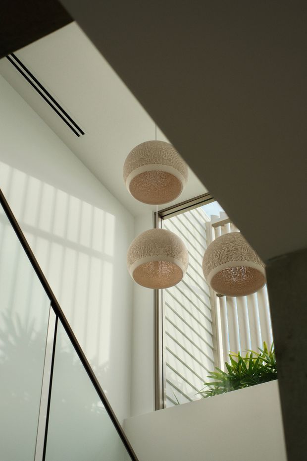 Sunlit contemporary interior corner featuring a tall glazed stairwell or landing with black-framed glass balustrading, a high white ceiling, three suspended woven spherical pendant lights, and a built-in planter with greenery. Strong geometric shadows and clerestory-style glazing create an airy vertical space.