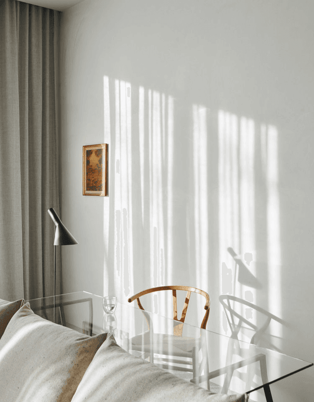 A softly lit minimalist interior corner with a glass dining table, curved wooden dining chairs, neutral upholstered seating in the foreground, full-height grey curtains, a slim black floor lamp, and a small framed artwork. Strong sunlight casts elongated window and furniture shadows across the white wall.