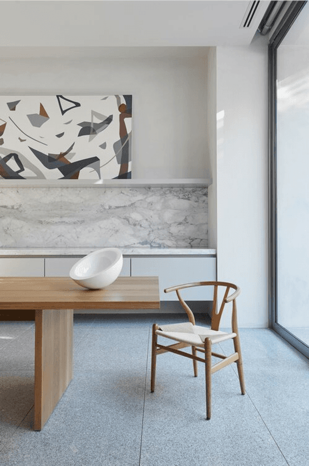 A refined contemporary dining area with a light timber dining table, sculptural white bowl, and wooden wishbone chair. The room features a large grey-and-white marble-look wall surface, pale built-in cabinetry, an abstract geometric artwork, polished concrete-like flooring, and full-height dark-framed glazing on the right. The palette is light, neutral, and softly contrasted, with natural timber accents.