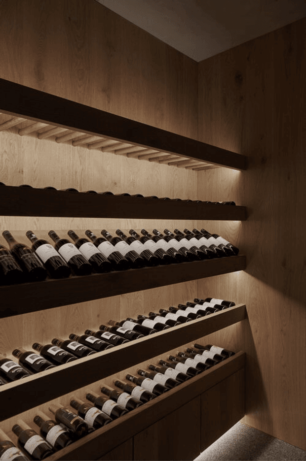 A dimly lit, built-in wine storage room lined with multiple dark timber shelves and angled bottle racks. Numerous wine bottles are displayed against warm wood-panelled walls, with concealed strip lighting illuminating the shelving. The space has a refined, bespoke, cellar-like appearance.