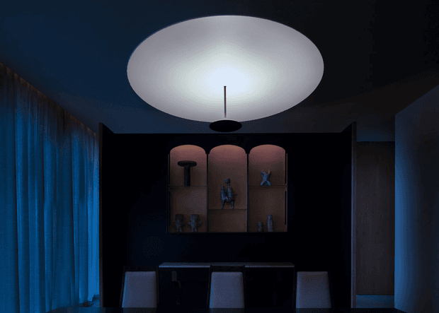 A dark, moody contemporary interior featuring a long dining or work table with several upholstered chairs, a dramatic oversized oval pendant light, and a black built-in display wall with rounded niches and small decorative objects. Floor-to-ceiling blue curtains line the left side, while dark walls and limited lighting create a theatrical atmosphere.