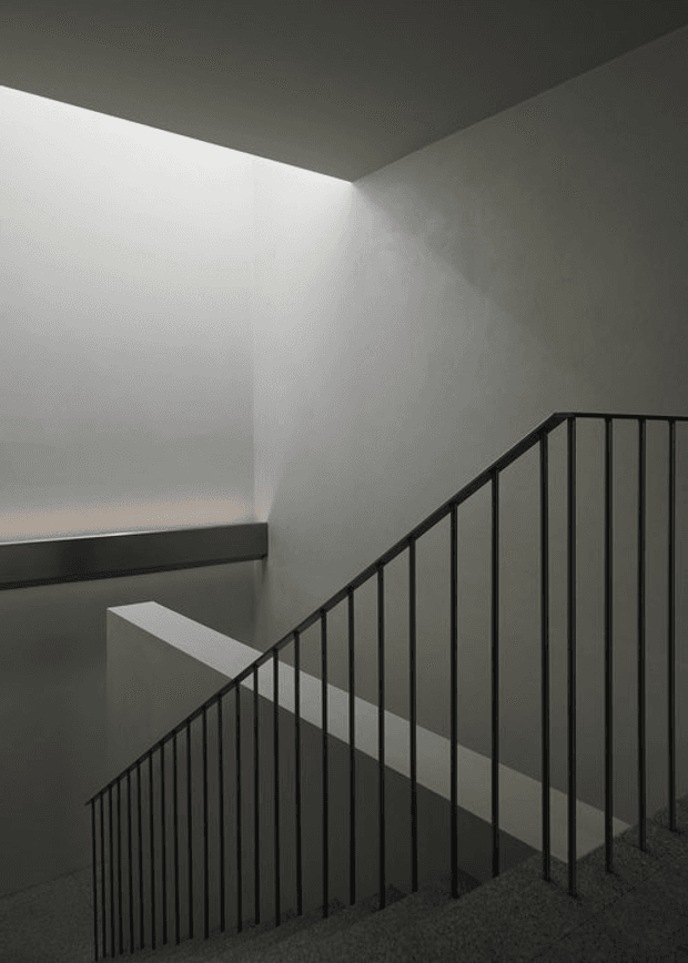 Minimalist interior stairwell viewed from an upper landing, featuring a dark metal balustrade with slender vertical pickets, grey carpeted steps, smooth pale grey plaster walls, and a strong skylight or clerestory-like opening washing the stairwell with diffuse light. The composition emphasizes clean geometric lines, shadow, and a subdued monochromatic palette.