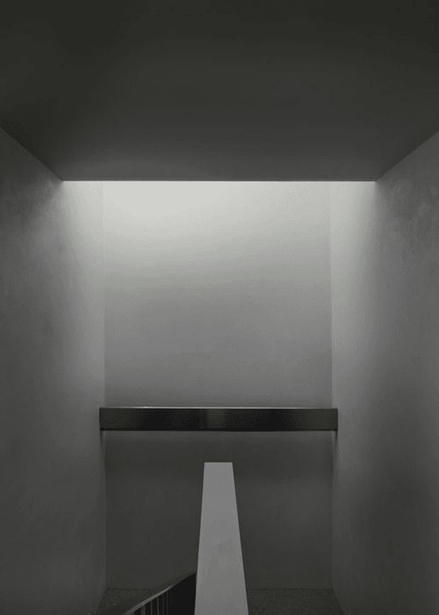 Minimalist architectural interior showing a narrow stairwell or stair landing enclosed by smooth grey walls. A dark horizontal structural beam spans the space, with a white tapered stair or balustrade element descending toward the foreground. The upper wall is softly illuminated, creating a stark monochromatic, moody composition.