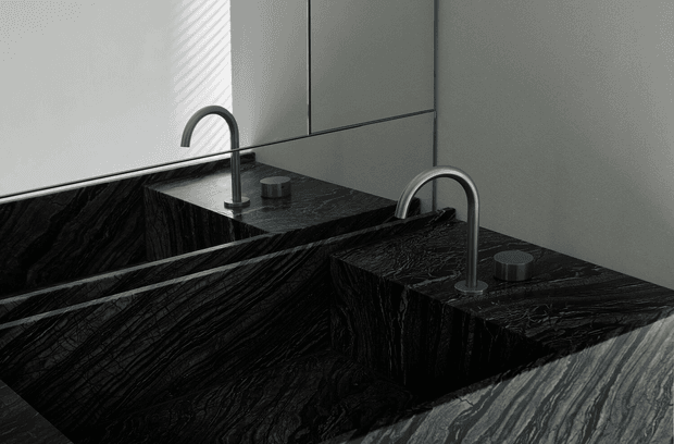 Close-up of a minimalist bathroom vanity with two integrated rectangular basins carved from dramatic black veined stone. Brushed metal gooseneck faucets and matching cylindrical controls sit on the stone surface, reflected in a large wall mirror. The composition is monochromatic, refined, and highly textural.