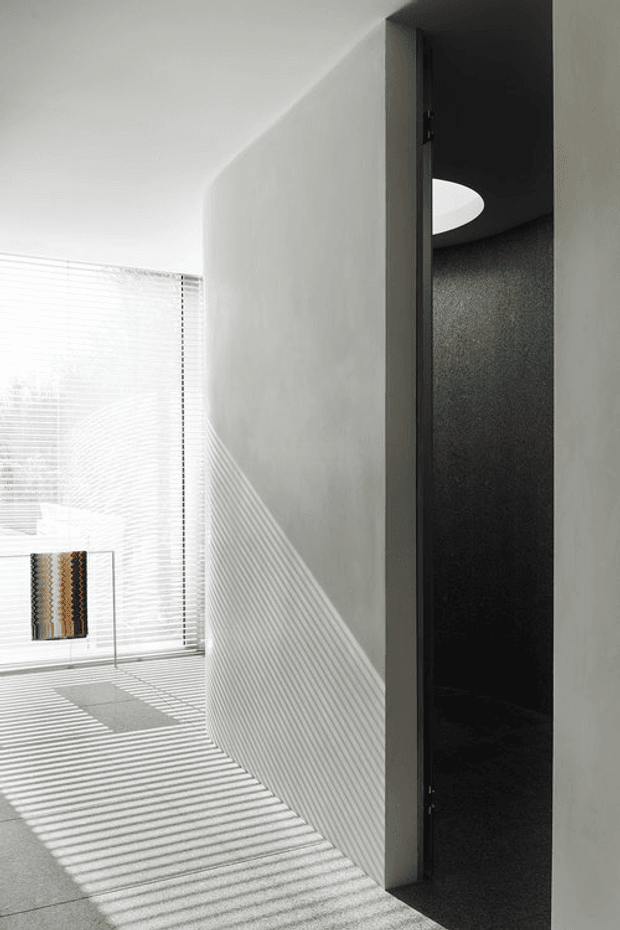A minimalist monochromatic interior hallway or passage with a curved white plaster wall, pale tiled floor, and strong linear sunlight filtered through venetian blinds. A dark recessed doorway opens into an adjoining room, with a circular ceiling light visible inside. The scene is high-contrast, quiet, and architectural, with a small framed textile or artwork near the bright window.