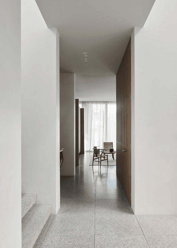 A minimalist interior view through a narrow hallway toward a bright sitting or dining area. White plaster walls and a high, clean-lined ceiling frame a warm timber feature wall and door on the right. Large pale terrazzo or stone-look floor tiles continue through the space, with a small dark wood dining table and chairs positioned near sheer floor-to-ceiling curtains. A short flight of matching terrazzo stairs is visible at the left foreground. The space is calm, refined, and sparsely furnished.