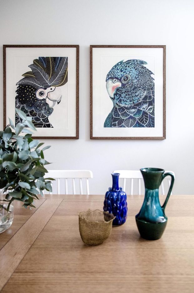 A bright dining area with a light timber dining table, white spindle-back chairs, and two framed illustrated bird artworks on a white wall. A leafy plant and blue-green ceramic vessels provide natural and jewel-toned accents.
