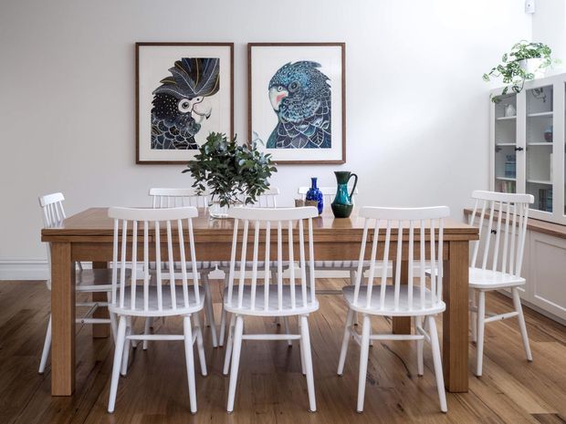 A bright, traditionally styled dining room centered on a large rectangular timber dining table surrounded by white spindle-back chairs. The room features wide timber floorboards, white walls and trim, a built-in glass-fronted display cabinet, framed bird artwork, and blue decorative ceramics on the table. Natural light creates an airy, welcoming atmosphere.